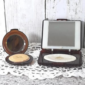 Op de afbeelding: Twee vintage compactpoeders. Eén is rond, open en bruin met een spiegel en een poederdons. De andere is rechthoekig, open en bruin met een spiegel en een poederdons. Beide liggen op een wit kanten oppervlak.