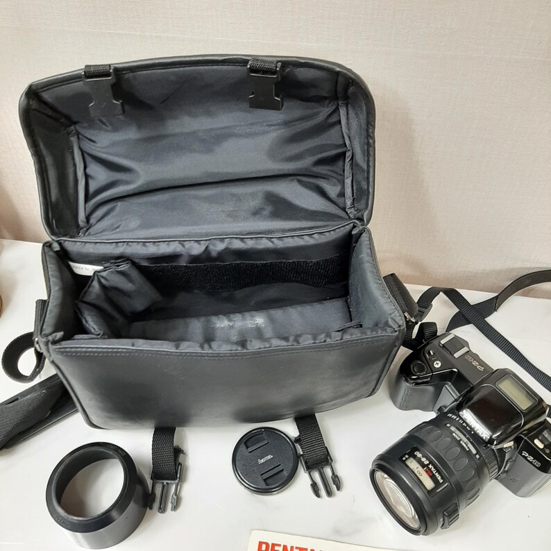 May include: A black camera bag with a zippered top and a shoulder strap. The bag is open and empty. A black camera lens with a silver ring is in the foreground. A black lens cap is on the table.