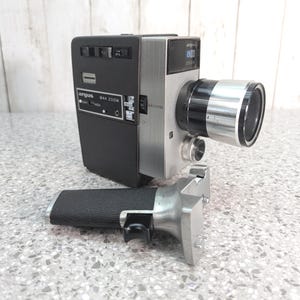May include: A vintage Argus M44 Zoom cine camera with a black body and silver metal front panel. The camera features a prominent lens, a side control panel with switches, and a detachable black textured pistol grip handle with a silver trigger.