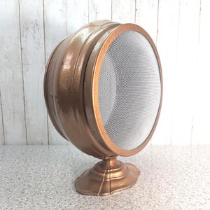 May include: A round, metallic copper-colored object with a wide, curved body and a circular front face covered by a fine silver-colored wire mesh screen. The item is supported by a decorative, pedestal-style base with a matching copper finish.