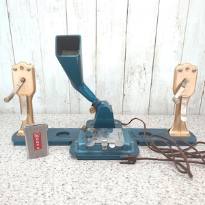 Peut inclure: Une étiqueteuse électrique bleue vintage avec un composant central en forme d'entonnoir, une base en métal et deux manivelles latérales dorées. Une petite étiquette rectangulaire argentée Kenco est fixée à l'avant, avec un cordon d'alimentation marron.