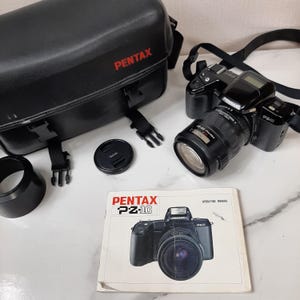 May include: A black Pentax PZ-10 camera with a lens attached, a black lens cap, a black camera bag with the word "PENTAX" in red lettering, and a black lens hood. The camera is sitting on a white surface. The camera has a black strap attached to it. The camera also has a black instruction manual with the words "PENTAX PZ-10" in white lettering.