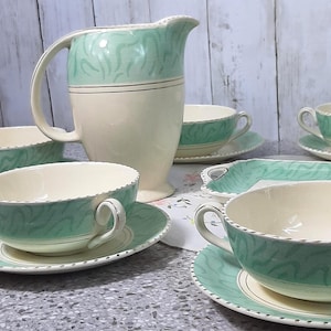 Puede incluir: Juego de té vintage con una jarra y tazas de té color crema con platillos a juego. El juego tiene un patrón de ondas verde menta en la parte superior de la jarra y las tazas. Los bordes están acentuados con un ribete plateado. Incluye una bandeja.