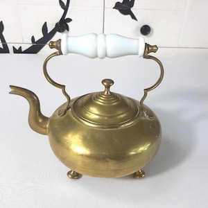 Può includere: Una teiera vintage in ottone con manico bianco e dettagli in ottone. La teiera ha un corpo arrotondato, un beccuccio curvo e poggia su tre piedini. Il coperchio è sormontato da un piccolo pomello in ottone.