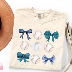 May include: Cream-colored t-shirt featuring watercolor baseballs and bows in shades of blue. The design includes baseballs with red stitching and various bow styles, creating a playful and sporty aesthetic.