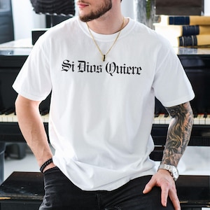 Comfort Colors t-shirt with “Si Dios Quiere” in dark Gothic Old English script — Spanish for “God willing.” Chicano-inspired lettering, vintage fade, soft cotton. Faith, family, y fe.