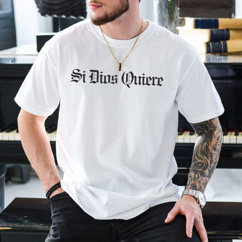 Comfort Colors t-shirt with “Si Dios Quiere” in dark Gothic Old English script — Spanish for “God willing.” Chicano-inspired lettering, vintage fade, soft cotton. Faith, family, y fe.