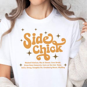 May include: White t-shirt with the text "Side Chick" in orange with stars. The text below reads "Mashed Potatoes, Mac & Cheese, Corn Bread, Green Bean Casserole, Corn on the Cob, Yams, Rolls, Gravy, Pumpkin Pie, Cranberry Sauce, Stuffing".
