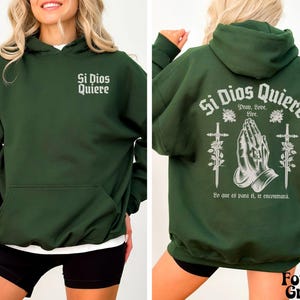 May include: A dark green hooded sweatshirt with the text "Si Dios Quiere" printed on the front and back. The back design includes a pair of hands in a praying position, two swords, and roses.