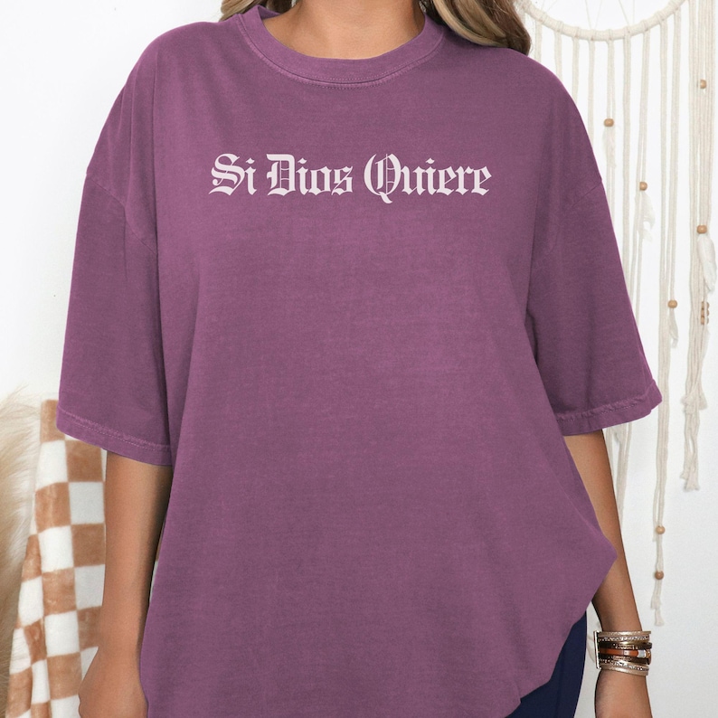 May include: A plum-colored short-sleeve t-shirt featuring the white text Si Dios Quiere printed in a gothic-style font across the chest. The garment has a crew neckline and a relaxed, loose-fitting silhouette with dropped shoulder seams.