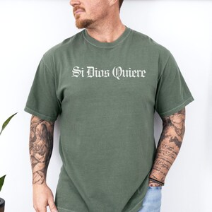 May include: A sage green short-sleeve t-shirt featuring the text Si Dios Quiere printed in a white gothic-style font across the chest. The garment has a crew neckline and a standard fit, constructed from a solid-colored fabric with short sleeves.