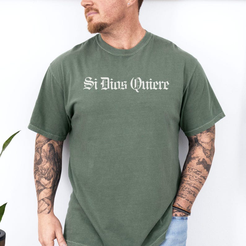 May include: A sage green short-sleeve t-shirt featuring the text Si Dios Quiere printed in a white gothic-style font across the chest. The garment has a crew neckline and a standard fit, constructed from a solid-colored fabric with short sleeves.