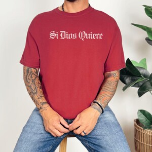 May include: A dark red short-sleeved t-shirt featuring the white text Si Dios Quiere printed in a gothic-style font across the chest. The garment has a crew neckline and a standard fit, providing a simple and clean look for casual wear.
