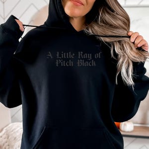 以下が含まれることがあります： ゴシック体の「A Little Ray of Pitch Black」の文字が入った黒いフード付きスウェットシャツ。スウェットシャツにはフロントポケットとドローストリングフードが付いています。生地は柔らかく、着心地の良い素材のようです。