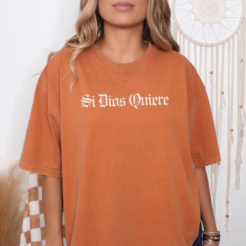 May include: A rust-colored, short-sleeved t-shirt featuring the text "Si Dios Quiere" printed in a white, gothic-style font across the chest. The garment has a crew neckline and a relaxed, loose-fitting silhouette with a solid, matte finish.