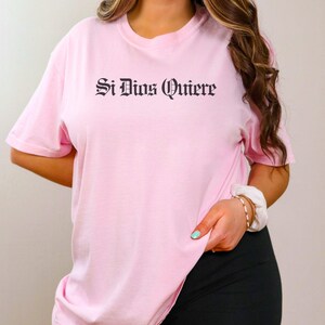 May include: A light pink short-sleeved t-shirt featuring the text Si Dios Quiere printed in a black gothic-style font across the chest. The garment has a crew neckline and a relaxed fit, providing a simple and clean look for casual wear.