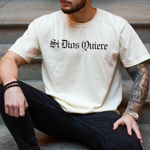 A Latino male model wearing a relaxed-fit heavy cotton Comfort Colors t-shirt featuring a black Old English Gothic font graphic that reads "Si Dios Quiere" across the front, styled in an urban city setting.