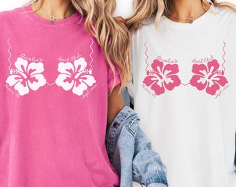 Può includere: Due magliette, una rosa e una bianca, con un design floreale. Il design include due fiori di ibisco stilizzati in rosa, con il testo "Amelia's Bach Club, Miami, Fl, Sept 2025."