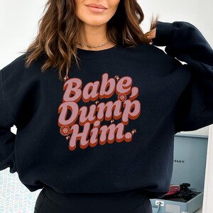 Puede incluir: Una sudadera negra con un gráfico de estilo retro que dice "Babe, Dump Him." El gráfico es de color rosa y rojo con un efecto brillante.