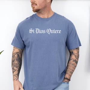 May include: A dusty blue short-sleeve crew neck t-shirt featuring the white text Si Dios Quiere printed in a gothic-style font across the chest. The garment has a standard fit with a ribbed neckline and a solid, muted blue color throughout.