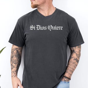 May include: A charcoal grey short-sleeved t-shirt featuring the text Si Dios Quiere printed in a white gothic-style font across the chest. The garment has a crew neckline and a standard fit, constructed from a solid-colored cotton fabric.