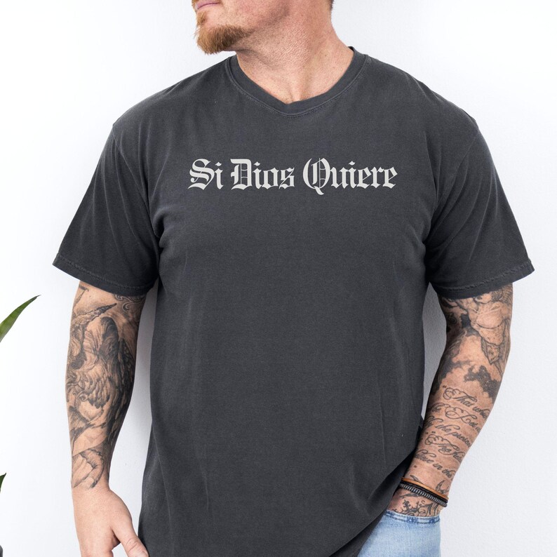 May include: A charcoal grey short-sleeved t-shirt featuring the text Si Dios Quiere printed in a white gothic-style font across the chest. The garment has a crew neckline and a standard fit, constructed from a solid-colored cotton fabric.