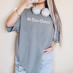 Close-up macro shot highlighting the premium textured fabric of a Comfort Colors shirt and the clean, detailed print of the phrase "Si Dios Quiere" in a striking Old English font.
