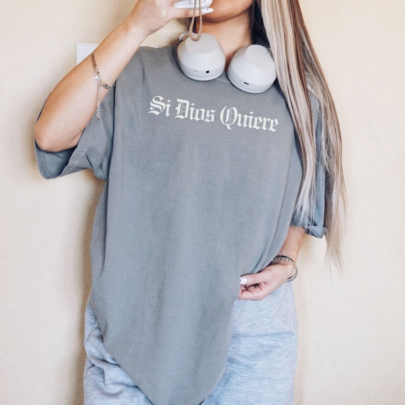 Close-up macro shot highlighting the premium textured fabric of a Comfort Colors shirt and the clean, detailed print of the phrase "Si Dios Quiere" in a striking Old English font.