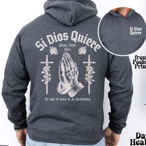 May include: Dark heather grey hoodie with a large graphic print on the back that says "Si Dios Quiere" with praying hands, roses, and swords. The front pocket has a smaller print that says "Si Dios Quiere".