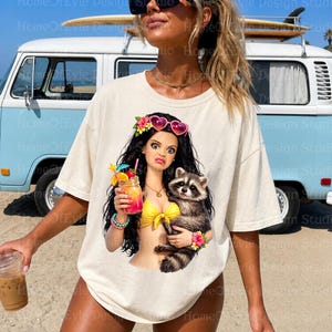 May include: A cream-colored short-sleeve t-shirt featuring a graphic of a cartoon character with long dark hair, heart-shaped sunglasses, and a yellow bikini top, holding a tropical drink and a raccoon, decorated with colorful hibiscus flowers.