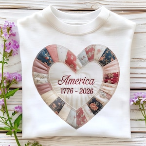 Puede incluir: Sudadera blanca con un diseño en forma de corazón. El corazón está hecho de parches de tela en varios colores y patrones, con la palabra "America" y los años "1776-2026" bordados en borgoña. Flores moradas a la izquierda y a la derecha.