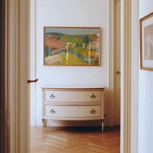 以下が含まれることがあります： 白壁に設置された、2つの引き出しと金色のトリムが付いたクリーム色のドレッサー。その上には、鮮やかな色の額入りの風景画が飾られています。部屋はクラシックでエレガントな雰囲気です。