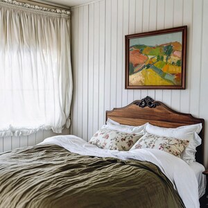 以下が含まれることがあります： オリーブグリーンのテクスチャードブランケット、白いシーツ、花柄のクッションで覆われたベッドのある寝室のインテリア。木製のヘッドボードと額入りの風景画が白いパネルの壁に掛けられています。透明な白いカーテンが左側にあります。