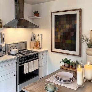 May include: A kitchen scene featuring a stainless steel stove and range hood. A framed abstract art piece hangs on the wall. A wooden table is set with plates, a teacup, and candles. Small potted plants add a touch of greenery.
