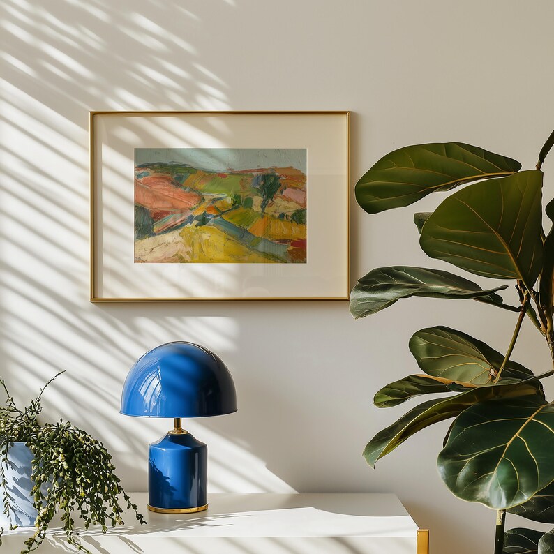 以下が含まれることがあります： 金色のフレームに入った風景画が、青いテーブルランプの上に飾られています。絵画は、赤、緑、黄色の色合いのカラフルな抽象的な風景を描いています。青いランプは丸いシェードと金色のベースです。植木鉢に入った植物が左側にあります。