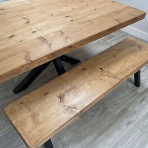 May include: A rectangular wooden dining table and a matching long bench, both featuring light brown natural wood grain surfaces with visible knots and sturdy black metal legs arranged in a cross-frame design for the table.