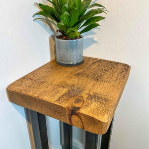 May include: A square wooden side table with a natural grain finish and a visible knot, supported by black metal legs. A green leafy plant sits in a light grey cylindrical pot on the surface of the table, which is positioned in a corner.