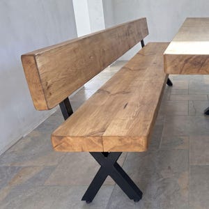 May include: A wooden bench with a backrest featuring a natural light brown wood grain finish and a thick seat. The bench is supported by black metal legs arranged in an X-shaped design, positioned next to a matching wooden table edge.