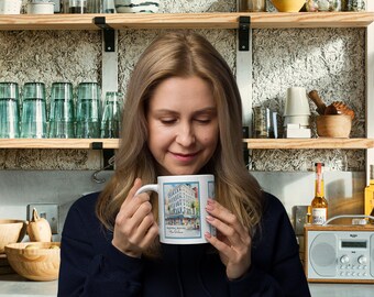 May include: A white ceramic mug with a colorful illustration of a building and the text "Broadway Market, New York City". The mug is being held by a person in a dark blue sweater.