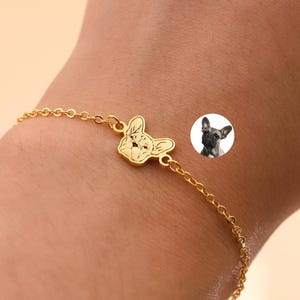 Puede incluir: Una pulsera dorada con un colgante de Bulldog Francés. El dije es una silueta detallada de la cara del perro. La pulsera se lleva en la muñeca, con una pequeña imagen de un Bulldog Francés en el fondo.