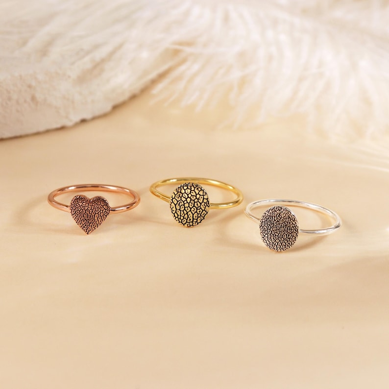 May include: Three rings with different designs. The first ring is rose gold and has a heart-shaped design. The second ring is gold and has a round design. The third ring is silver and has a round design.