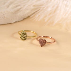 May include: Two simple gold rings with textured designs. One ring has a round design and the other has a heart shape.