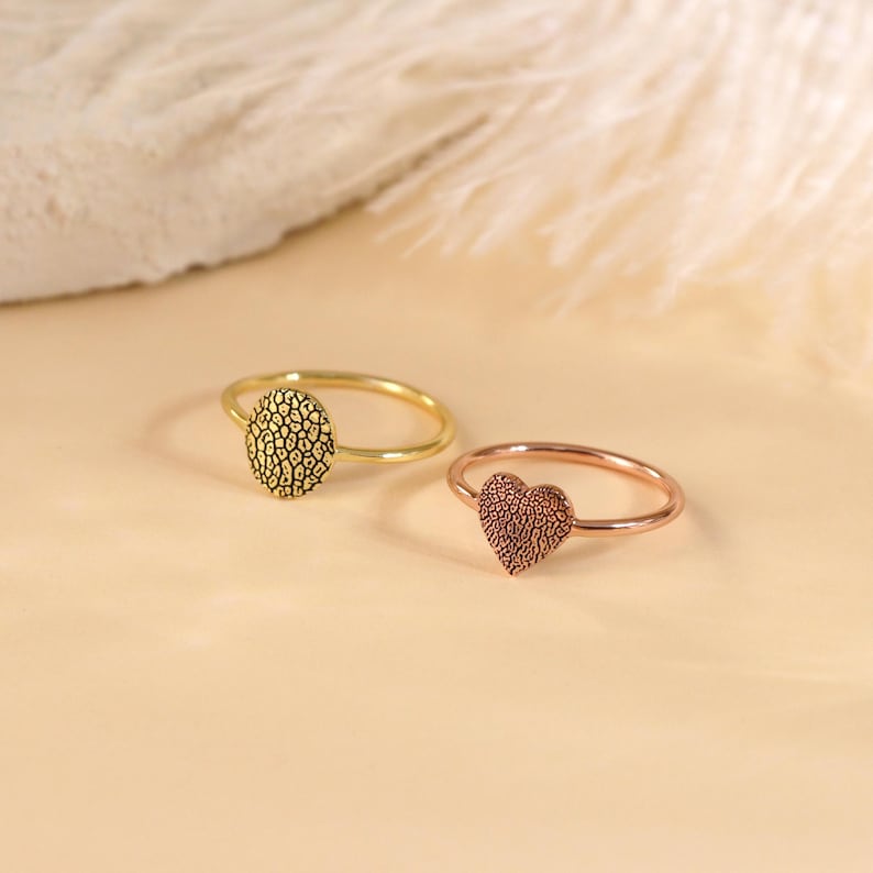 May include: Two simple gold rings with textured designs. One ring has a round design and the other has a heart shape.
