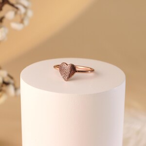 May include: A rose gold heart-shaped ring with a textured surface. The ring is displayed on a white pedestal.