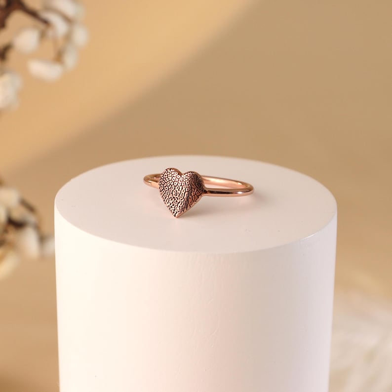 May include: A rose gold heart-shaped ring with a textured surface. The ring is displayed on a white pedestal.