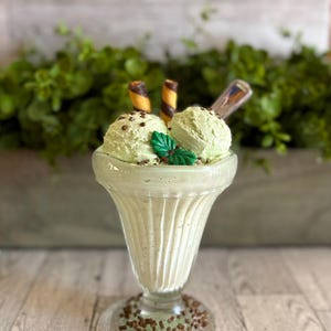 May include: A glass sundae dish filled with two scoops of green mint ice cream topped with chocolate sprinkles and two chocolate wafer sticks. A sprig of mint sits on top of the ice cream.