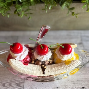 Pu&ograve; includere: Un banana split sundae con tre palle di gelato, guarnito con panna montata, ciliegie e salsa al cioccolato. Il sundae viene servito in una ciotola di vetro con una banana in cima.