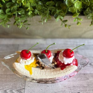 Pu&ograve; includere: Un dessert di banana split con tre palle di gelato guarnite con panna montata e ciliegie. Il gelato &egrave; giallo, marrone e rosa. Il dessert &egrave; in una ciotola di vetro trasparente.