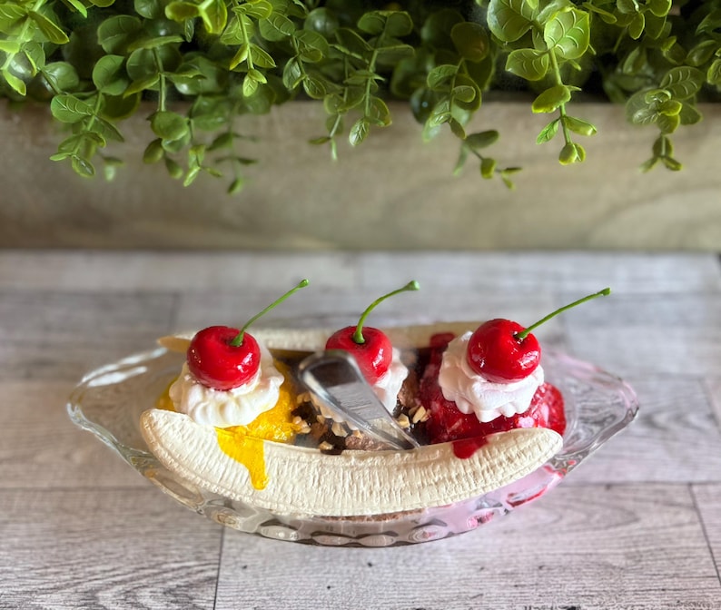 Pu&ograve; includere: Un dessert di banana split con tre palle di gelato guarnite con panna montata e ciliegie. Il gelato &egrave; giallo, marrone e rosa. Il dessert &egrave; in una ciotola di vetro trasparente.