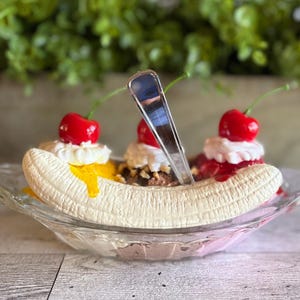 Pu&ograve; includere: Un banana split sundae con tre palle di gelato, guarnito con panna montata, ciliegie e un cucchiaio. Il banana split &egrave; in una ciotola di vetro.